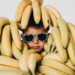 Young Boy in Sunglasses Sticking His Head Through a Pile of Bananas