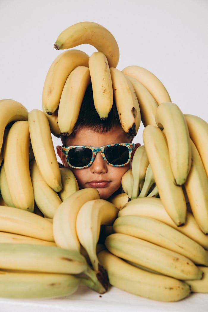 Young Boy in Sunglasses Sticking His Head Through a Pile of Bananas 